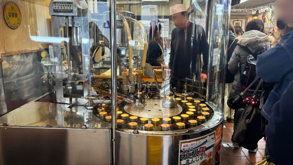 箱根湯本の食べ歩きで人気の温泉まんじゅうを店頭で焼いている様子と、購入を待つ観光客の風景。
