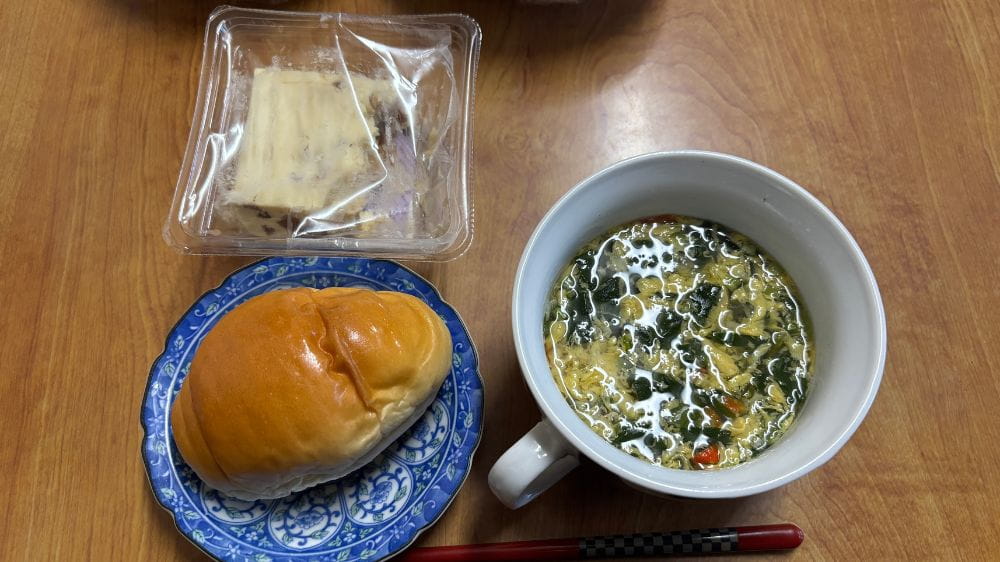 ロールパンと具だくさんのスープ、パック入りのおかずを並べたシンプルな食事。