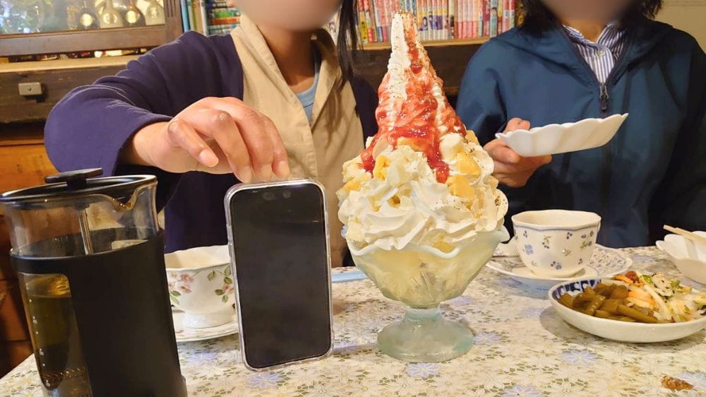 喫茶のんのん名物の巨大なチーズパフェ。スマートフォンと比べても大きさが際立つ、ボリューム満点のメニュー。