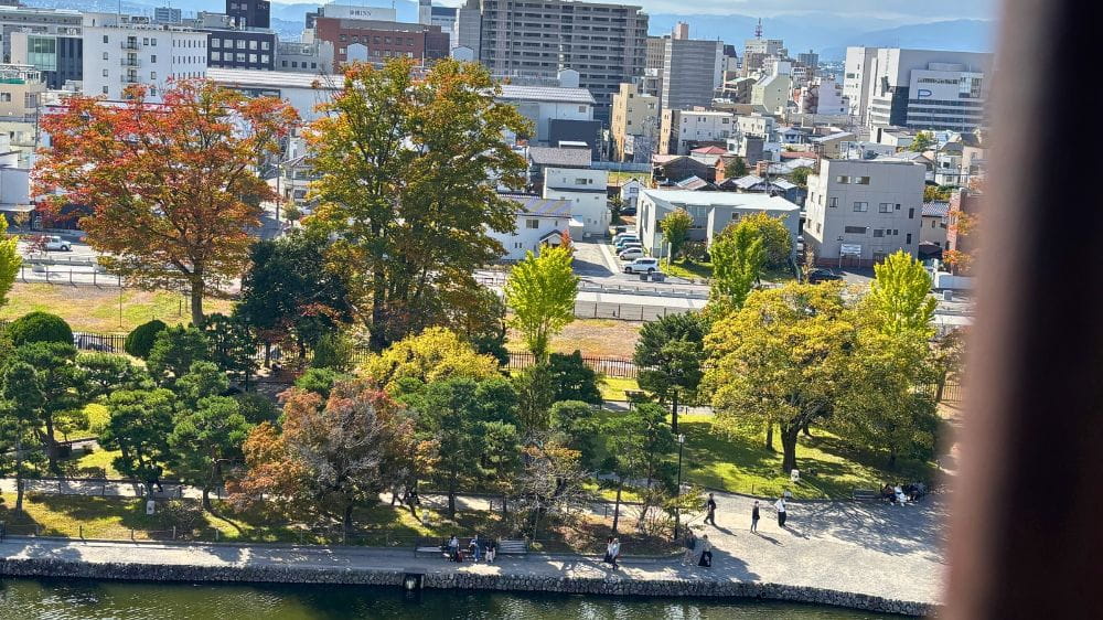 松本城天守の最上階の窓から見た、堀沿いの色づき始めた木々と松本市街の建物