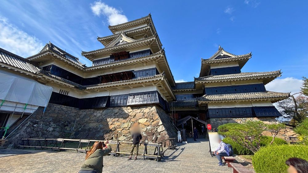 青空の下にそびえる国宝松本城の天守と石垣、見学者が行き交う入口付近の様子